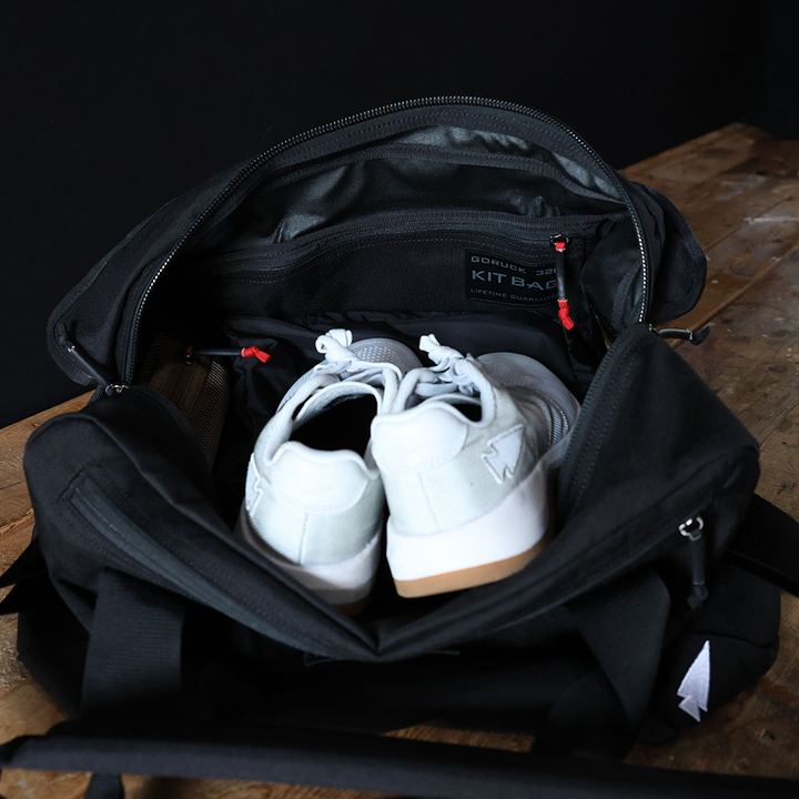 Open black GORUCK Kit Bag with a pair of white sneakers inside, resting on a wooden surface.