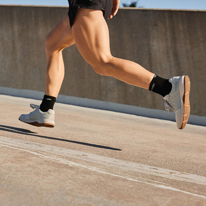 A person running on a concrete surface is wearing black shorts and socks, complemented by gray Ballistic Trainers from GORUCK, in the Lunar Rock and Gum colorway with a Silver Reflective Spearhead. These shoes are crafted from durable CORDURA® Ballistic Nylon material to endure any level of intensity during functional fitness workouts.