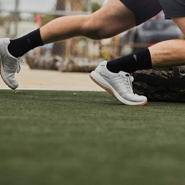 A person wearing GORUCK Men's Ballistic Trainers in Lunar Rock + Gum with a Silver Reflective Spearhead and black socks is running on a green sports field, dragging a weighted sled behind them. The background features a blurred outdoor setting with a chain-link fence and parked vehicle.