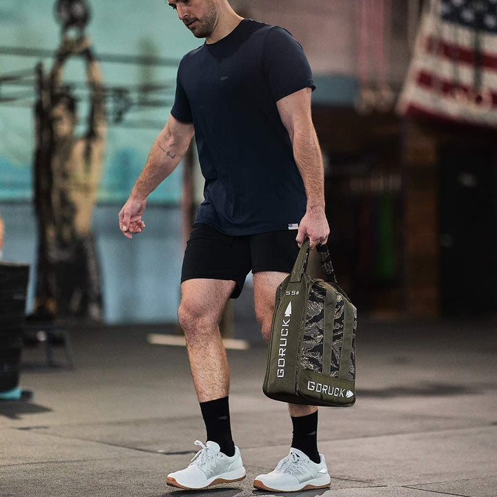 A person in athletic gear walks in a gym carrying a camo-patterned GORUCK bag made of durable CORDURA Ballistic Nylon. They wear a navy shirt, black shorts, and GORUCK Men's Ballistic Trainers in Lunar Rock with Gum soles and a Silver Reflective Spearhead. An American flag and gym equipment are visible in the background.