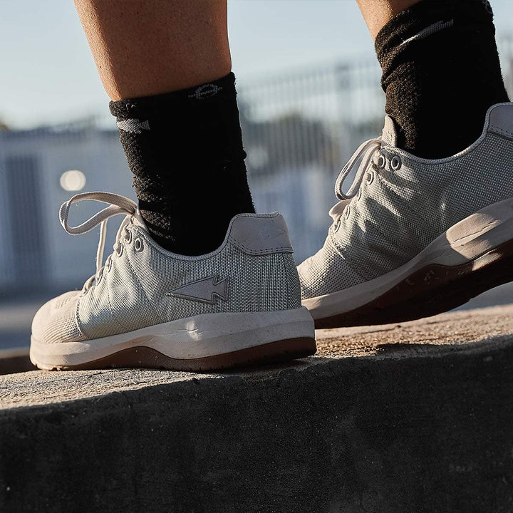 A close-up shows someone wearing GORUCK's Men's Ballistic Trainers in Lunar Rock + Gum, featuring a silver reflective spearhead design. These athletic shoes are crafted from durable CORDURA Ballistic Nylon for exceptional wear, and the person is standing on a concrete ledge in black socks with a blurred fence in the background—ideal functional fitness footwear.