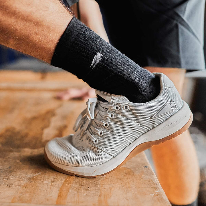 A close-up shot captures someone wearing a black sock paired with GORUCK's Men's Ballistic Trainers in Lunar Rock and Gum, enhanced with a Silver Reflective Spearhead. The sneaker, crafted from CORDURA Ballistic Nylon, rests on a wooden surface featuring gum soles and 3X Support for superior stability, tightly laced for maximum performance.