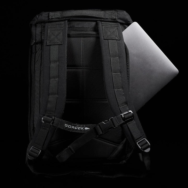 A contemporary M23 - Cordura rucksack by GORUCK, designed with robust straps and a discreet logo, holds a laptop partially tucked inside against a dark backdrop. This rucksack is constructed from durable materials to ensure long-lasting quality and style.