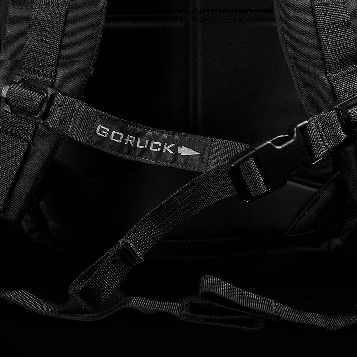 Close-up of a black M23 - Cordura rucksack showcasing the distinct GORUCK strap and buckle. The image emphasizes its modern, tactical design featuring durable materials, reinforced stitching, and adjustable straps set against a dark background.