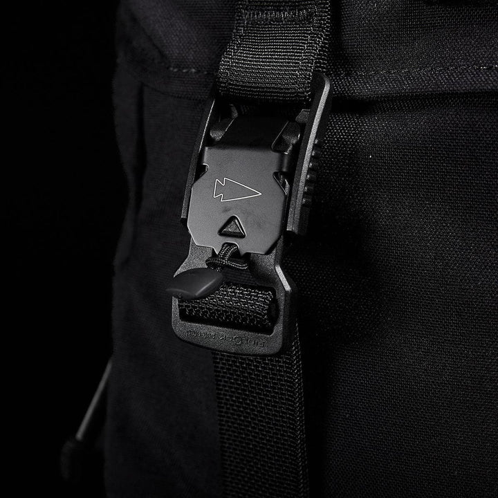 Close-up of a black buckle with an arrow symbol, fastened on a strap against the sleek Cordura fabric of the M23 rucksack by GORUCK. The buckle boasts a modern design, adding a subtle accent to the durable materials.