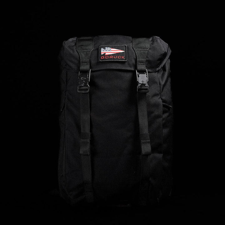 A modern rucksack is displayed against a dark background. This black M23 - Cordura rucksack features two buckle straps and a patch with an American flag design alongside the brand name GORUCK on the upper front, crafted from durable materials for lasting use.