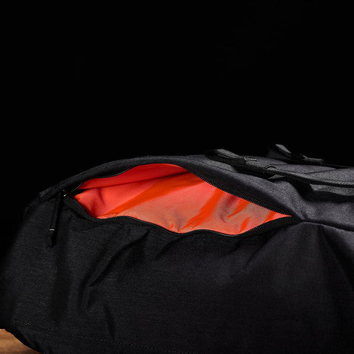 A contemporary GORUCK M23 - Cordura rucksack with an open side pocket reveals its vibrant orange interior against a dark backdrop. The zipper of the M23 is partially unzipped, highlighting the contrasting colors crafted from durable materials.