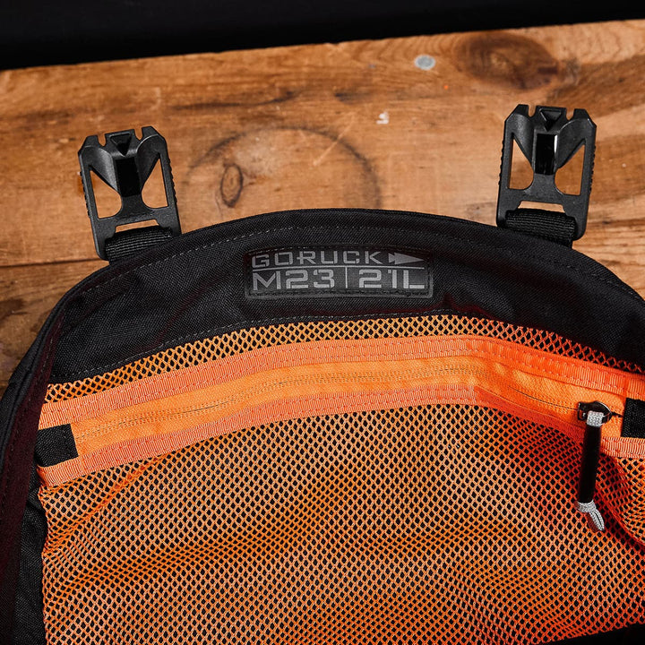 A close-up of an open black backpack labeled GORUCK M23 - Cordura against a wooden background highlights its durable materials. The interior, featuring orange mesh pockets and two black clips at the top, exemplifies the functionality of a modern rucksack.