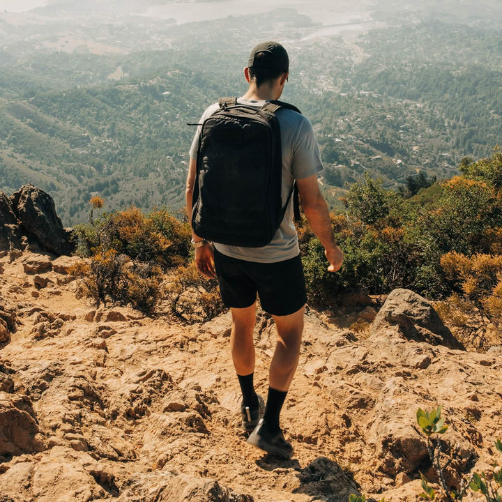 A man with a GORUCK Mackall backpack and Black + Forged Iron trail shoes, designed with a triple compound outsole for extra grip, hiking downhill on a rocky mountain trail with a scenic valley below.