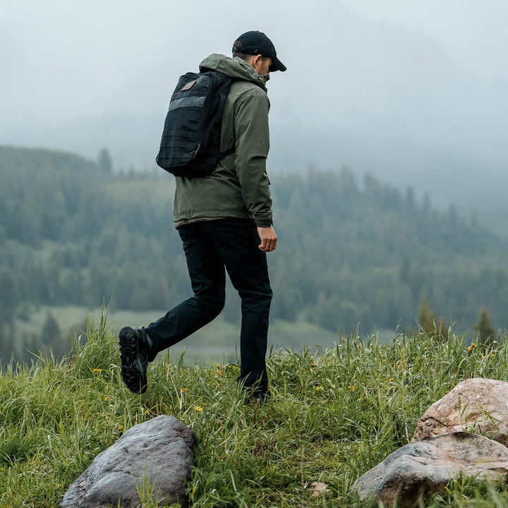 A hiker on a grassy hill in foggy weather, wearing a green jacket and backpack, confidently strides with GORUCK's Mackall shoes in Black + Forged Iron, featuring a triple compound outsole for superior grip.