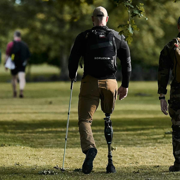 A person with a prosthetic leg and walking stick strolls through a park, sporting a GORUCK backpack and Mackall - Black + Forged Iron rucking shoes.