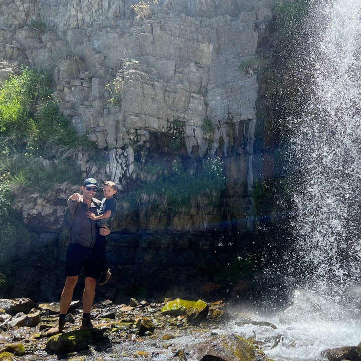 A person holding a child near a waterfall with a rocky cliff background, both wearing GORUCK's Merino Challenge Socks, which promise comfort and durability.