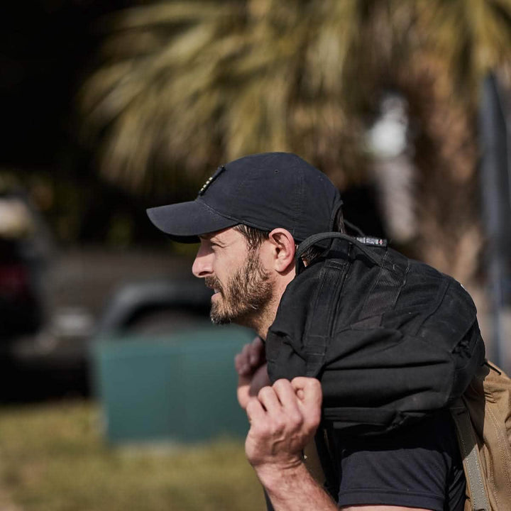 A man wearing a Performance TAC Hat - TOUGHDRY by GORUCK carries a black backpack over his shoulder. He is outdoors, with palm trees and a park-like setting in the background.