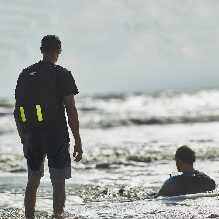 A person stands on the beach wearing a black t-shirt, Men’s Challenge Shorts by GORUCK with cargo pockets made from Lightweight ToughDry® fabric, and a backpack. Another sits in the water facing the ocean. The scene is calm with gentle waves and a cloudy sky.