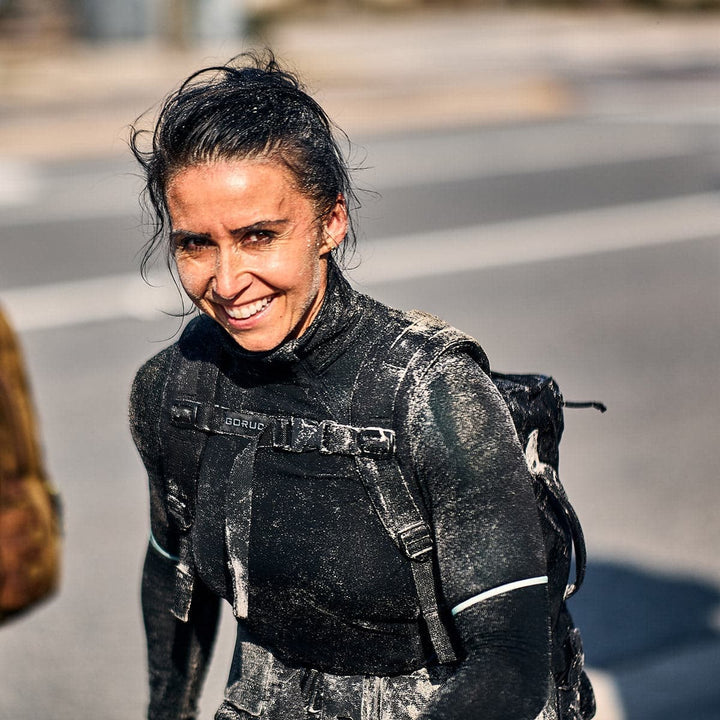 A smiling woman with wet hair and a muddy wetsuit stands on the street, her GORUCK backpack featuring the Training Sternum Strap and MOLLE webbing hinting at her adventurous spirit.