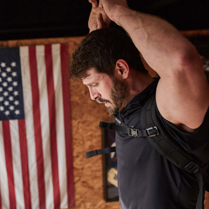 A man with a determined demeanor stretches in front of a wall decorated with an American flag, gearing up for the day ahead. His GORUCK equipment features MOLLE webbing and the Training Sternum Strap, ensuring he's prepared for any challenge while maximizing ease of movement.