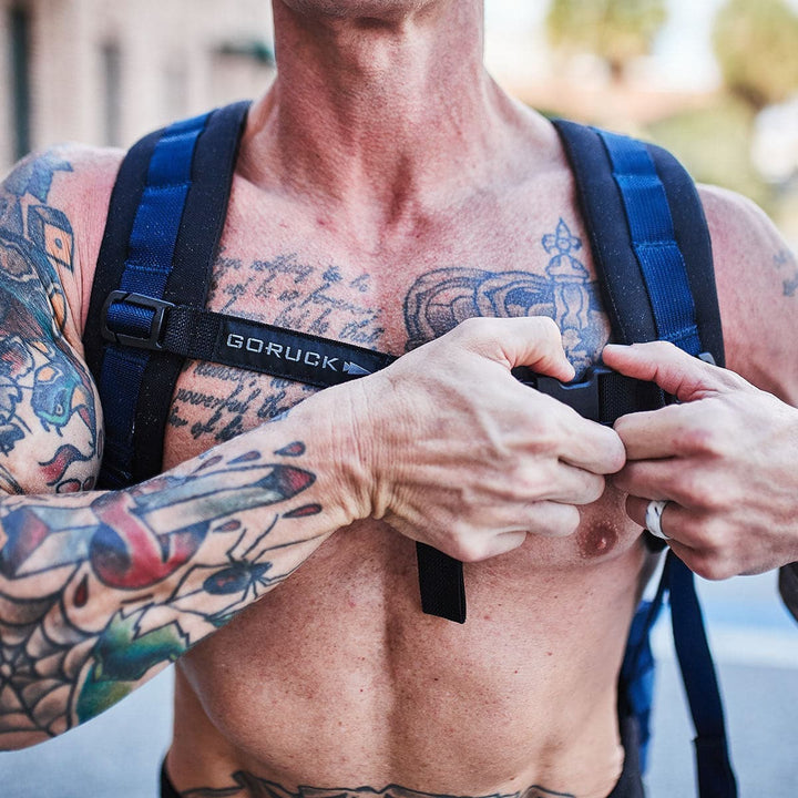 A tattooed person stands outdoors, adjusting the GORUCK Training Sternum Strap on their backpack for optimal weight distribution.