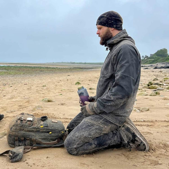 A person kneels on a sandy beach, wearing a hat and rugged clothing made from abrasion-resistant fabric. Holding a container, their Men’s Challenge Pants - Lightweight ToughDry® by GORUCK are ideal for the setting. A backpack rests beside them against the backdrop of a cloudy sky and expansive shoreline.