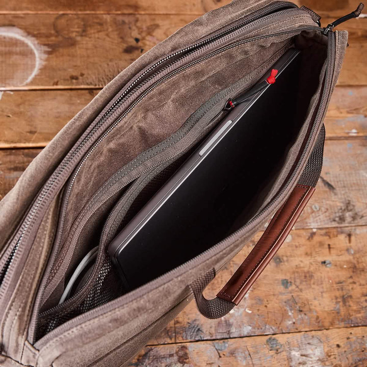 A brown waxed canvas shoulder bag from GORUCK lies open on a wooden table, with a laptop partially inside. The bag includes a zipper closure, asymmetrical straps, and a handle accented with leather.