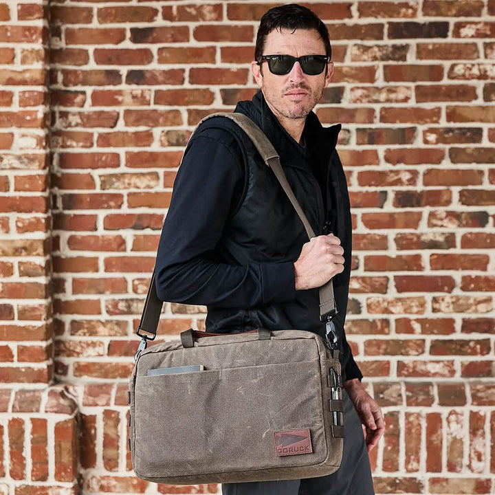 A man wearing sunglasses and a dark jacket holds the GORUCK Shoulder Bag - Waxed Canvas over his shoulder. He stands in front of a brick wall, looking towards the camera with an air of confidence.