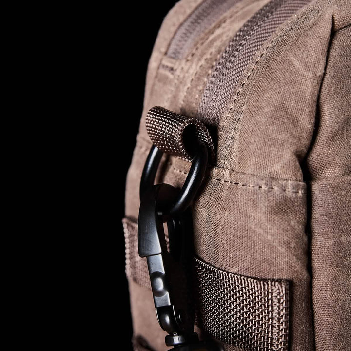 Close-up of a GORUCK Shoulder Bag - Waxed Canvas in brown, featuring a black carabiner attached to a loop, perfectly suited for professional settings. The textured waxed canvas and visible stitching add to its elegance. A black background accentuates the bag's sophisticated details, offering options for MOLLE customization.