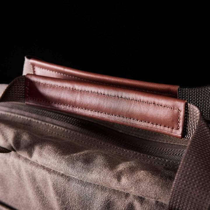 Close-up of a brown leather strap connected to the waxed canvas fabric of the GORUCK Shoulder Bag. The photo emphasizes the stitching detail on the leather handle, set against a dark background, highlighting its sophisticated design.