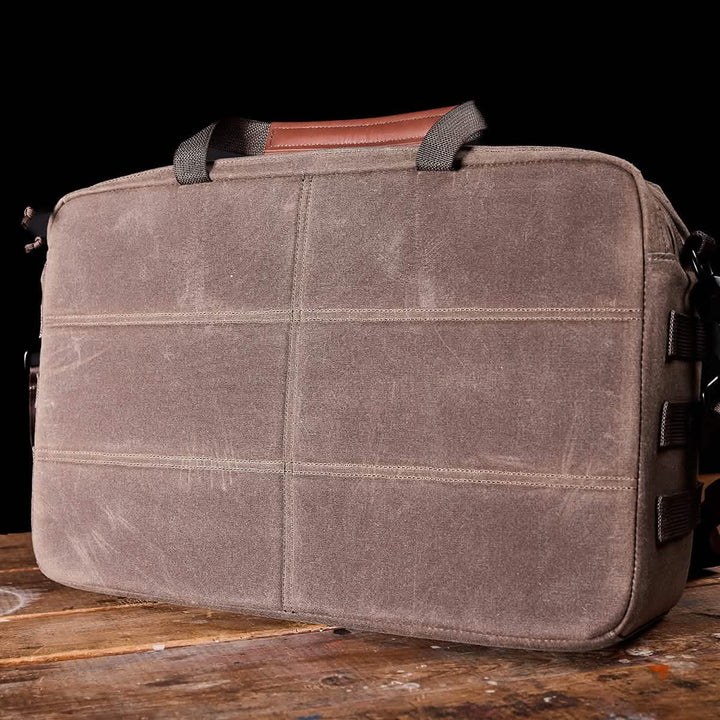 A GORUCK Shoulder Bag - Waxed Canvas, featuring a warm brown tone with complementary black handles, rests gracefully on a wooden table. Its classic style is accentuated by the sleek black background, making it an ideal blend of timeless design and modern functionality for the discerning professional.