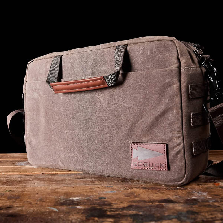 A brown GORUCK Shoulder Bag - Waxed Canvas rests on a wooden surface against a black background. This professional messenger bag showcases a leather patch with the brand logo, multiple straps, and a sturdy handle, while offering MOLLE customization for enhanced versatility.