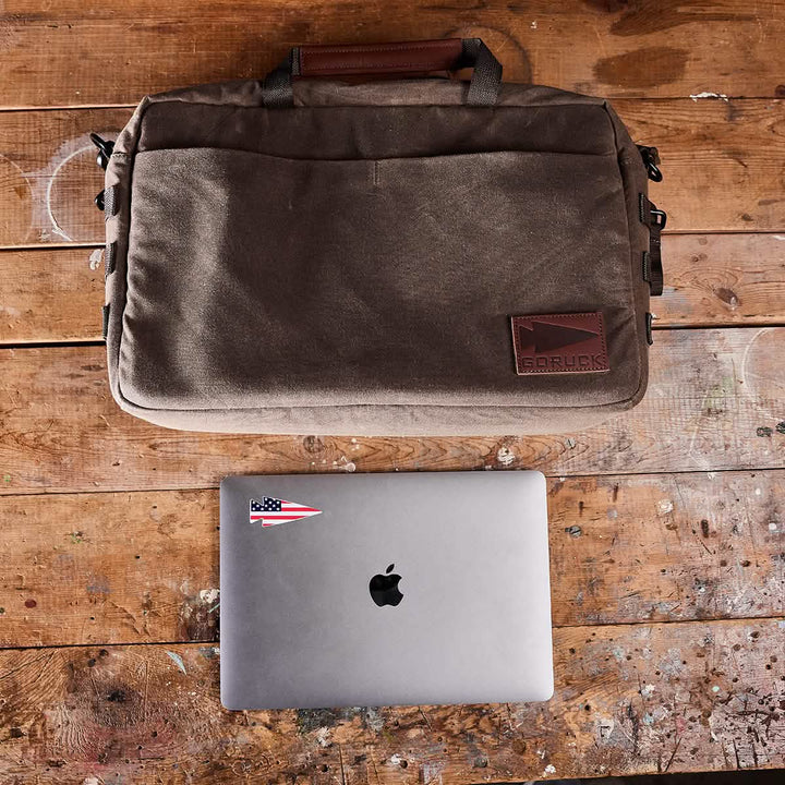 A brown waxed canvas shoulder bag from GORUCK is resting on a worn wooden table, above a closed silver MacBook adorned with a small sailboat sticker. The bag features MOLLE customization, a leather handle, and sports a patch on the front. Paint splatters can be seen on the surface of the table.