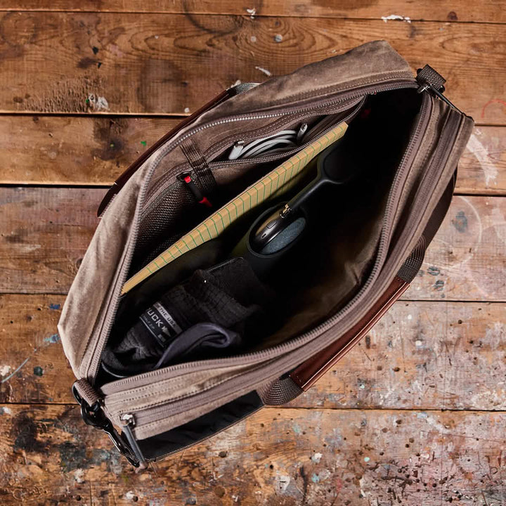 An overhead view of an open GORUCK Shoulder Bag - Waxed Canvas on a rustic wooden surface showcases its professional design. Inside, items like a notepad, pen, pair of socks, and a black object are neatly arranged. The bag features a leather handle, multiple pockets, and suggests MOLLE customization capabilities.