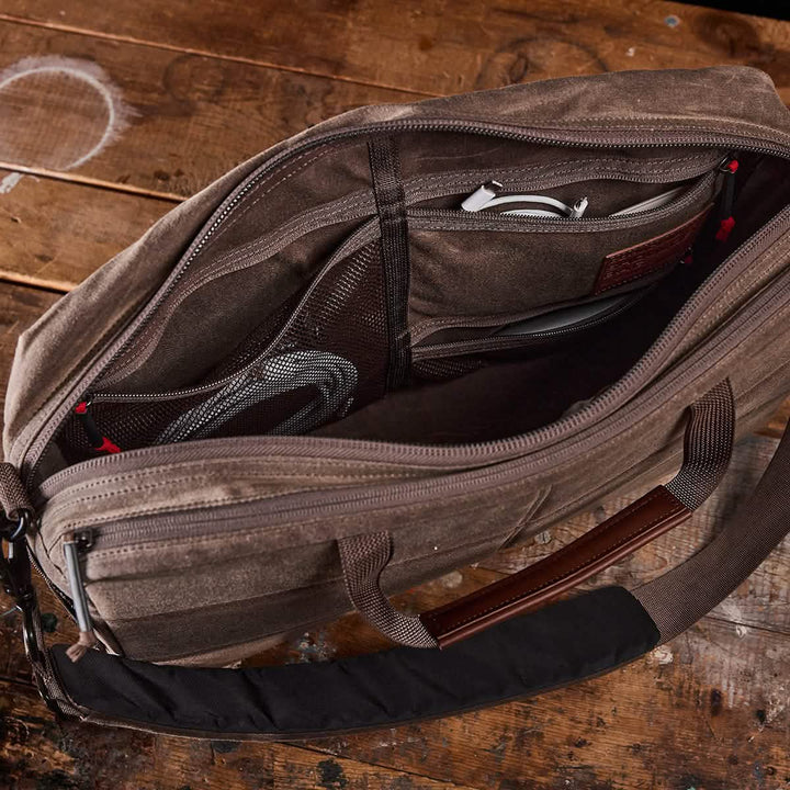 Showcase the GORUCK Shoulder Bag - Waxed Canvas, a professional brown canvas design with an asymmetrical style on a wooden surface. It features multiple compartments, including a mesh pocket, leather patch, and zipper pouch. The bag also boasts padded shoulder straps for added comfort.