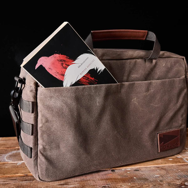 A waxed canvas shoulder bag by GORUCK rests on a wooden table, with a copy of Mexican Gothic peeking from its outer pocket. This brown messenger bag features a leather handle, MOLLE customization, and a patch with the GORUCK logo against the black backdrop, making it ideal for any discerning professional.