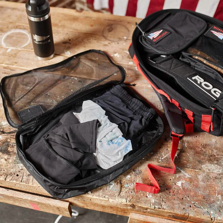 An open backpack on a wooden table features a mesh compartment containing folded clothing and a cloth, all neatly organized with GORUCK Packing Cubes. A water bottle rests nearby, while part of an American flag waves gently in the background. These packing cubes are your perfect travel companion.