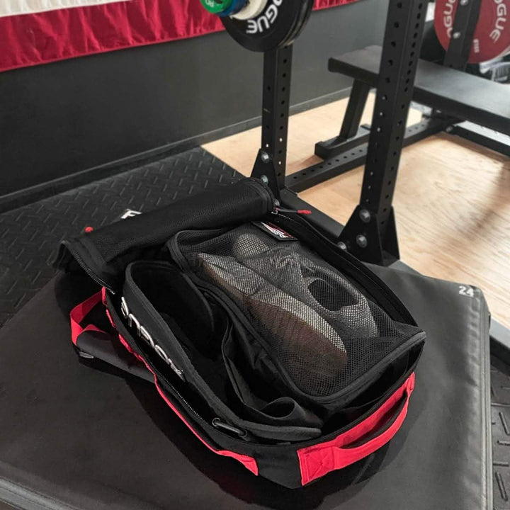 A GORUCK gym bag designed from 200D nylon, featuring mesh compartments, sits open on a weight bench with a pair of sneakers peeking out. In the background, a portion of a barbell setup can be seen alongside a red and white flag adorning the wall.