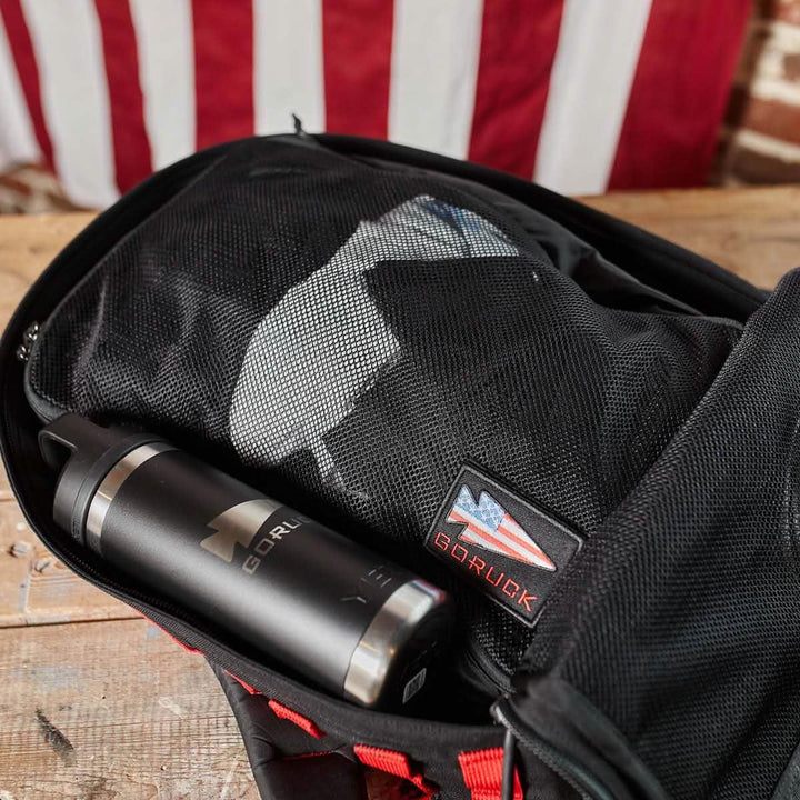 A travel ruck is open, displaying a GORUCK water bottle and a mesh pouch with items neatly organized inside, likely with the help of GORUCK Packing Cubes. An American flag patch adorns the 200D nylon bag, while a blurred flag lies in the background on a wooden surface.