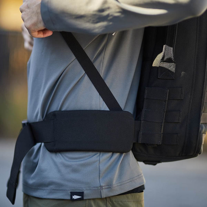 A person, wearing a gray long-sleeve shirt and green pants, adjusts the strap on a black GORUCK CORDURA rucksack featuring a Padded Hip Belt.