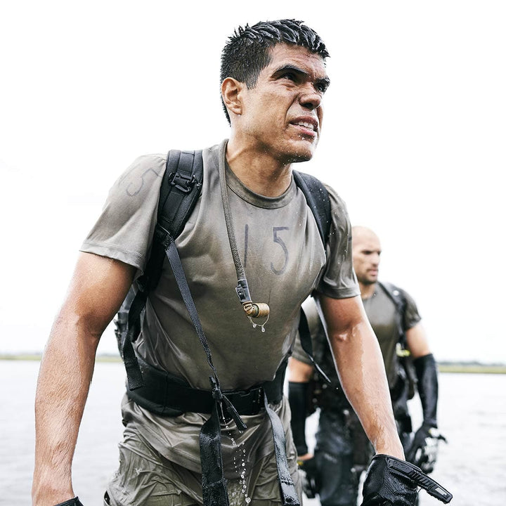 The man in wet gear, with a robust GORUCK backpack equipped with a Padded Hip Belt, strides determinedly forward. Behind him, another person in similar gear follows suit, their packs securely fastened for the rugged journey ahead.