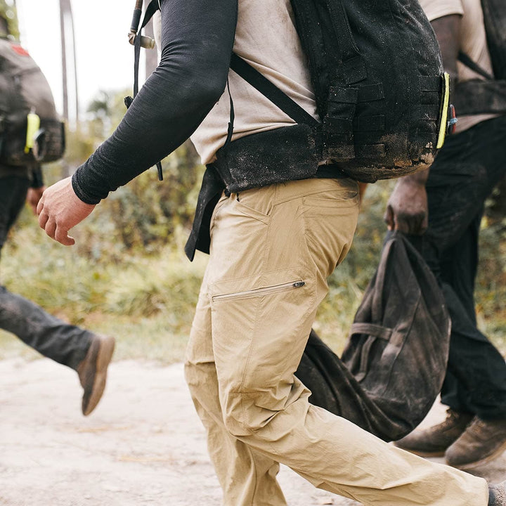 A group of hikers navigates a dirt path surrounded by lush grass, their backpacks showcasing the durable CORDURA fabric and enhanced comfort with the GORUCK Padded Hip Belt.