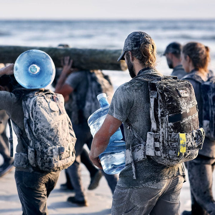 A group of people, with GORUCK Padded Hip Belts securely fastened around their waists and rucksacks on their backs, are covered in mud as they carry large jugs of water and logs while walking along a sandy beach.