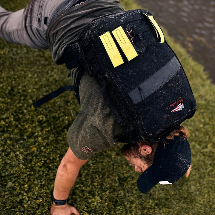 A person is doing a push-up on the grass, wearing a cap and showcasing a GORUCK backpack featuring durable yellow CORDURA straps.