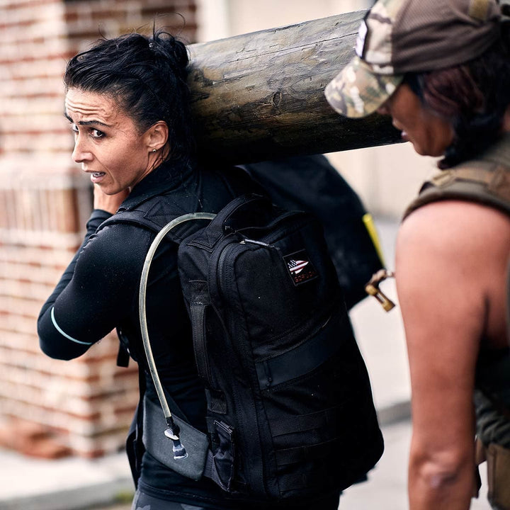 Two people carrying a large log; one looks back, confident with a GORUCK hydration backpack equipped with the Padded Hip Belt for added comfort. Made from durable CORDURA fabric, their gear guarantees resilience and ease during the heavy ruck through rugged trails.