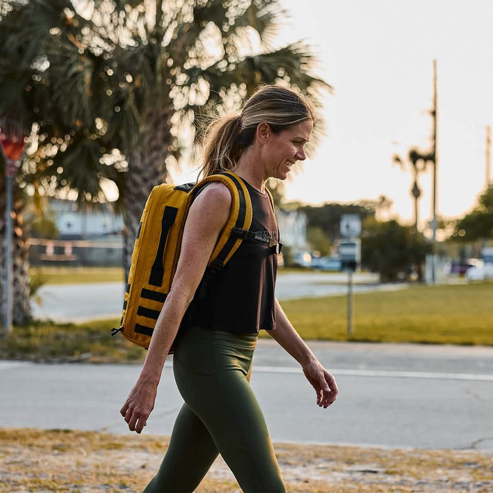 A woman strolls outdoors wearing a yellow backpack, the GORUCK Women's Performance Tank crafted from ToughMesh™ fabric in black, and green leggings. She seems joyful under the sunlight, with trees and a street behind her, evoking the tranquility of a walk near the Italian Alps.