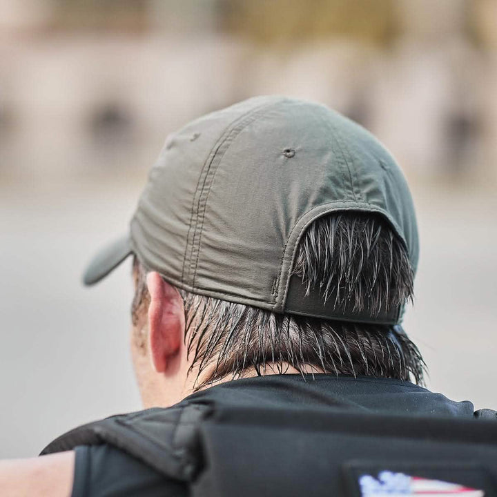 A man wearing a GORUCK Performance TAC Hat - TOUGHDRY, in green, reveals wet hair visible from behind as he stands outdoors in a black vest.