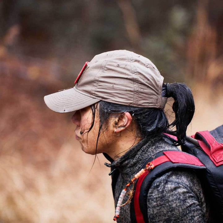 A person wearing the GORUCK Performance TAC Hat - TOUGHDRY and a jacket hikes through the forest, carrying a backpack.