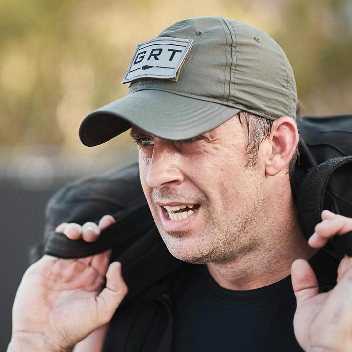 A man wearing a green Performance TAC Hat by GORUCK appears determined, carrying a black bag with VELCRO® straps on his shoulder outdoors.