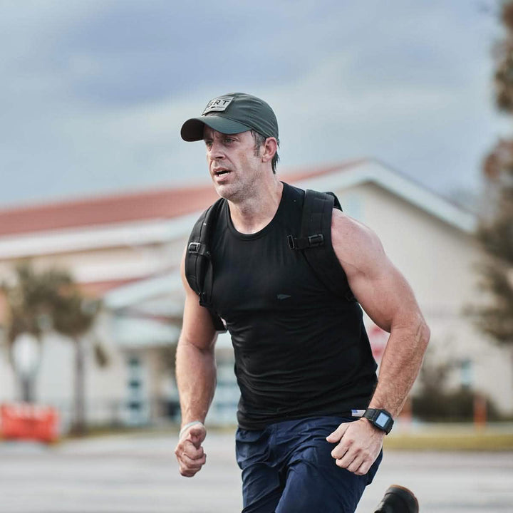 A man in a black tank top and a GORUCK Performance TAC Hat featuring TOUGHDRY fabric is running outdoors with a backpack and smartwatch. His outfit ensures he stays comfortable and cool with its sweat-wicking technology.