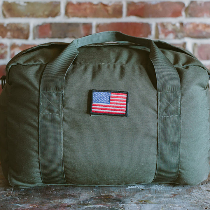 The GORUCK Kit Bag, in an olive green hue and featuring an American flag patch, evokes the design of a Special Forces Flight Bag and is displayed on a stone surface with a brick wall backdrop.