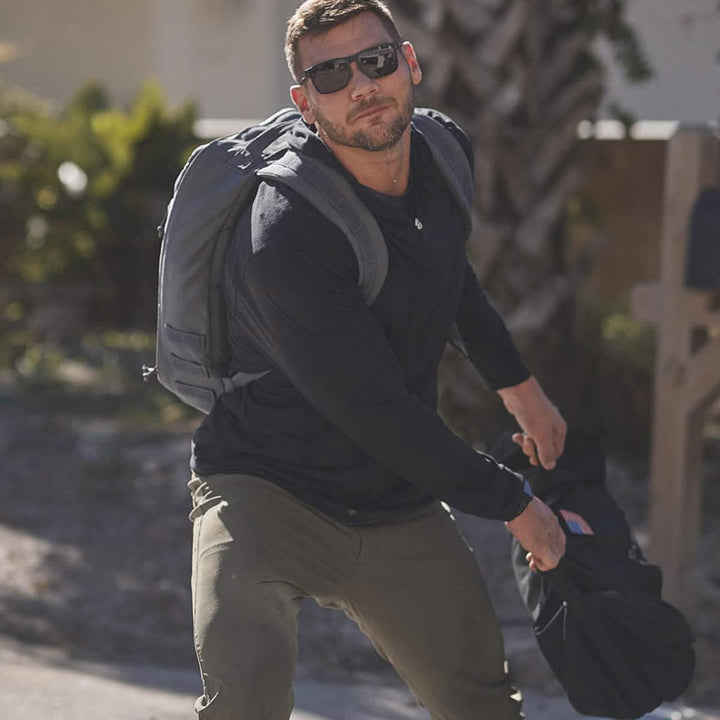 A man wearing sunglasses and a backpack is crouching on a sunny day. He's dressed in a black long-sleeve shirt and GORUCK’s olive green Men’s Simple Pants made with Midweight ToughDry® material, holding onto a black jacket. The background features greenery and a blurred outdoor setting.