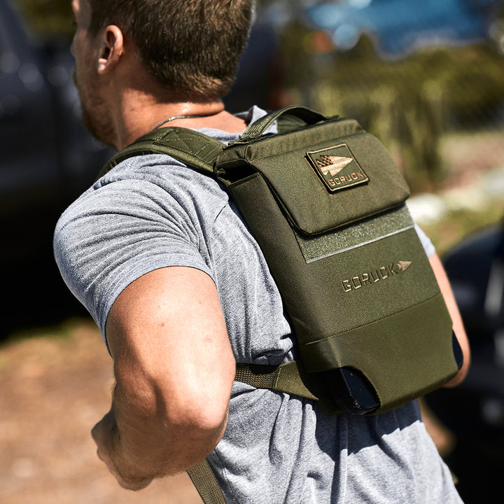 A person is spotted from behind wearing a green GORUCK Ruck Plate Carrier 3.0 over a gray t-shirt while outdoors, showcasing its ergonomic lumbar support that ensures comfort on any adventure.