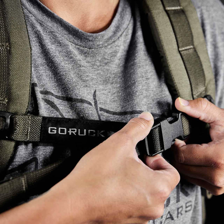 A person wearing a gray t-shirt deftly adjusts the GORUCK Training Sternum Strap on an olive backpack featuring MOLLE webbing.